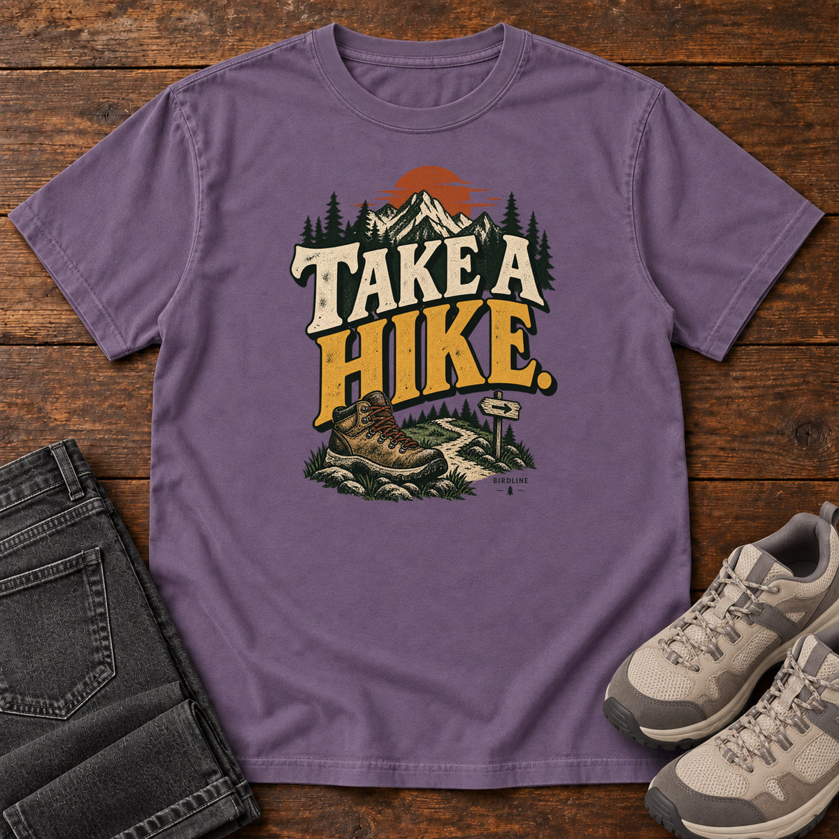 Grape Take a Hike T-Shirt with a hiking boot, mountain trail and orange sun around bold cream and gold lettering; front flat lay on wood.