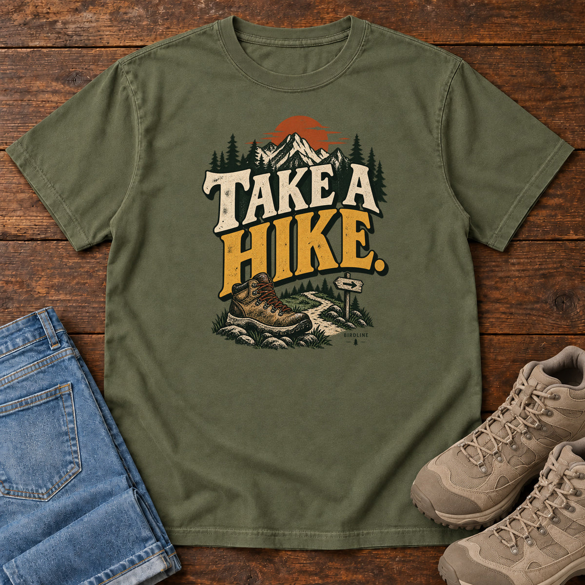 Hemp Take a Hike T-Shirt with a hiking boot, mountain trail and orange sun around bold cream and gold lettering; front flat lay on wood.