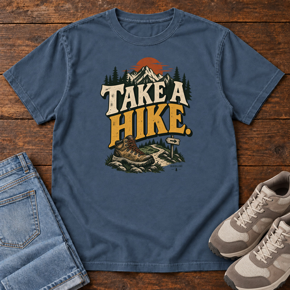 Midnight Take a Hike T-Shirt with a hiking boot, mountain trail and orange sun around bold cream and gold lettering; front flat lay on wood.