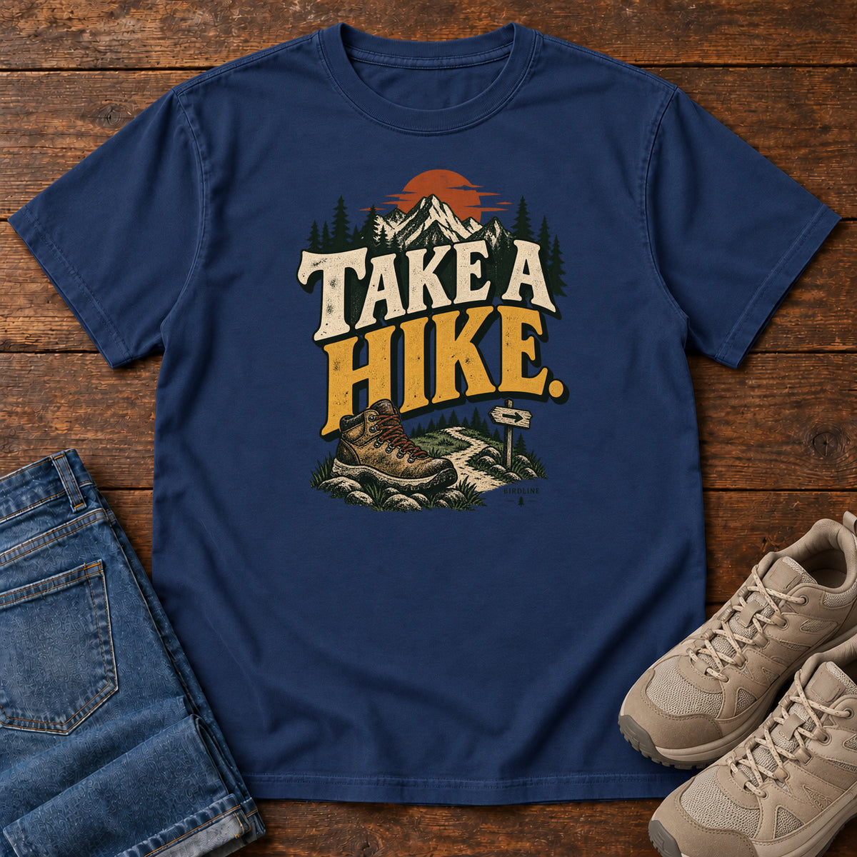 True Navy Take a Hike T-Shirt with a hiking boot, mountain trail and orange sun around bold cream and gold lettering; front flat lay on wood.