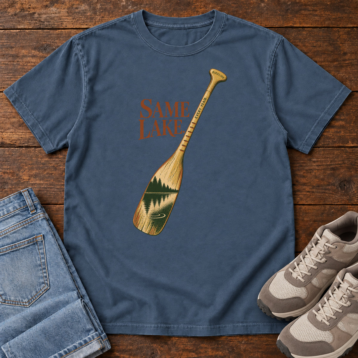 Midnight Same Lake Every Year T-shirt with a paddle and lake illustration, laid flat on a wooden surface.