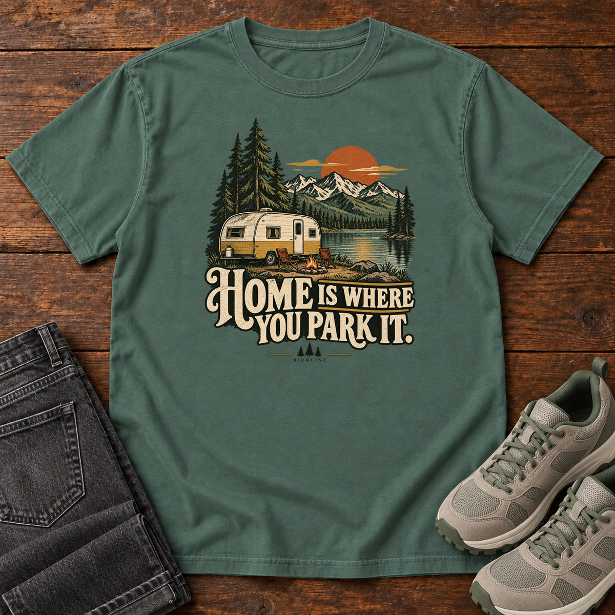 Home Is Where You Park It T-Shirt in Blue Spruce, front laid flat on wood, featuring a cream camper beside a mountain lake, pine trees and an orange sun above slogan lettering.