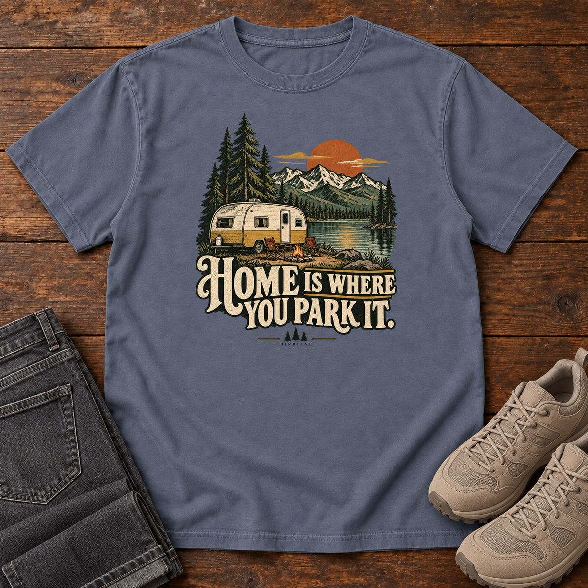 Home Is Where You Park It T-Shirt in Denim, front laid flat on wood, featuring a cream camper beside a mountain lake, pine trees and an orange sun above slogan lettering.