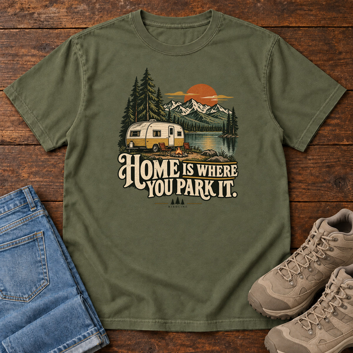 Home Is Where You Park It T-Shirt in Hemp, front laid flat on wood, featuring a cream camper beside a mountain lake, pine trees and an orange sun above slogan lettering.