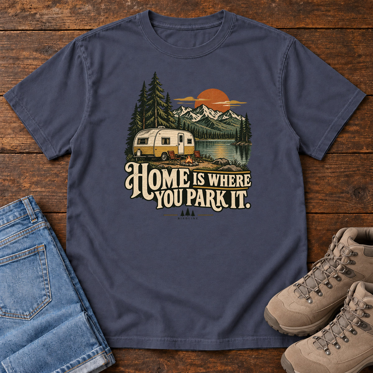 Home Is Where You Park It T-Shirt in Navy, front laid flat on wood, featuring a cream camper beside a mountain lake, pine trees and an orange sun above slogan lettering.