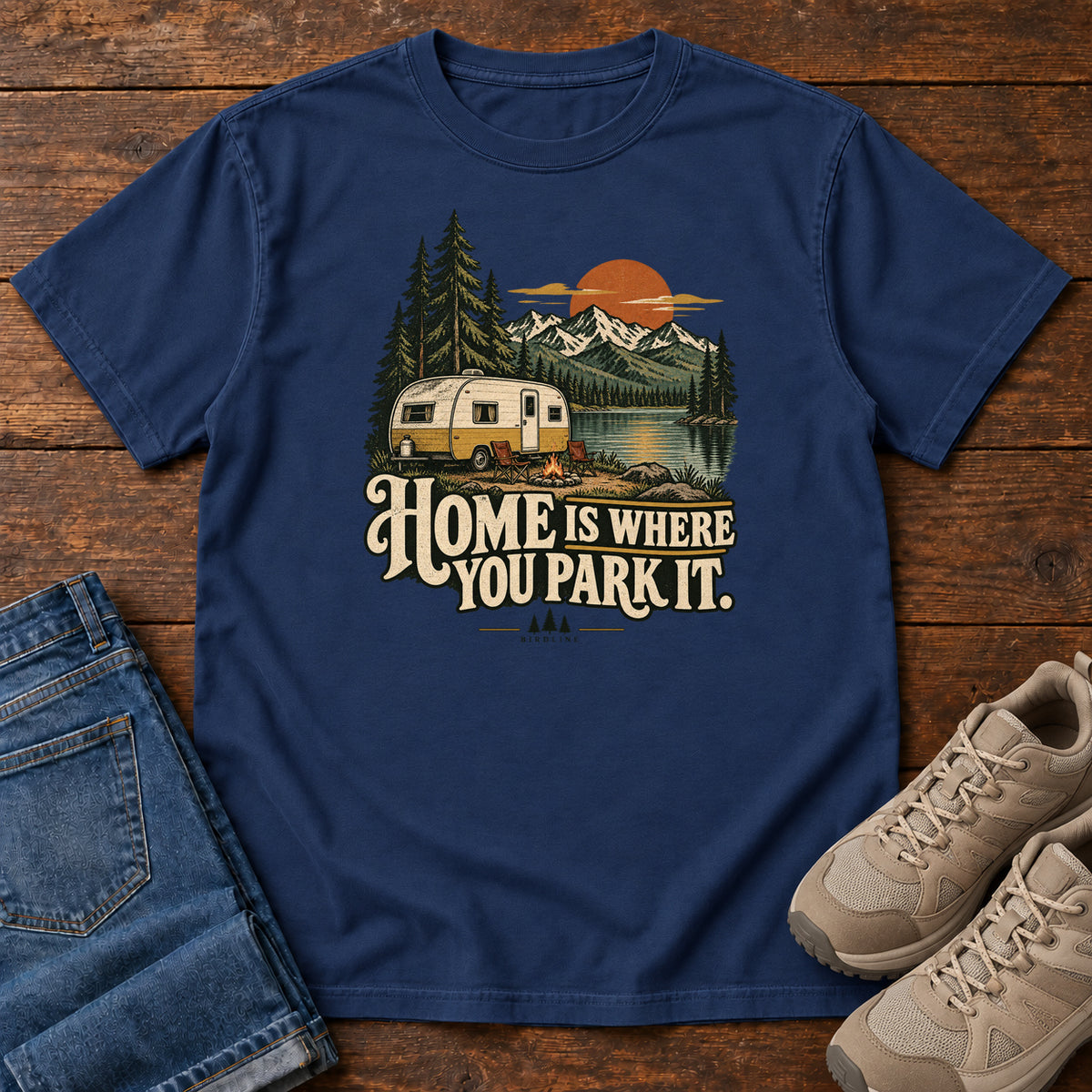 Home Is Where You Park It T-Shirt in True Navy, front laid flat on wood, featuring a cream camper beside a mountain lake, pine trees and an orange sun above slogan lettering.