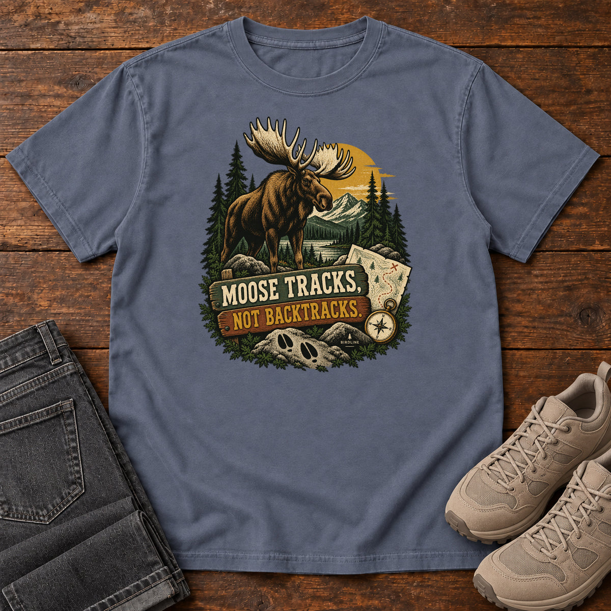 Denim Moose Tracks Not Backtracks T-Shirt with a moose in a mountain forest above a map, compass and cream and gold lettering, front flat lay on wood.