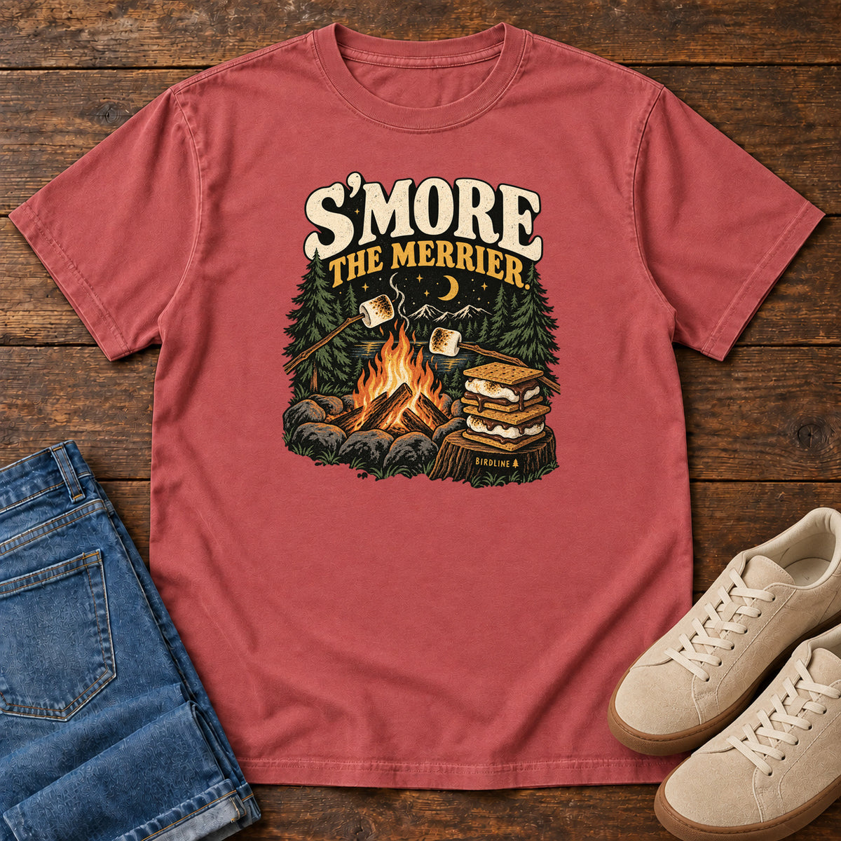 Crimson Smore the Merrier T-Shirt with a campfire, marshmallows on sticks and stacked s’mores beneath a crescent moon; front flat lay on wood.