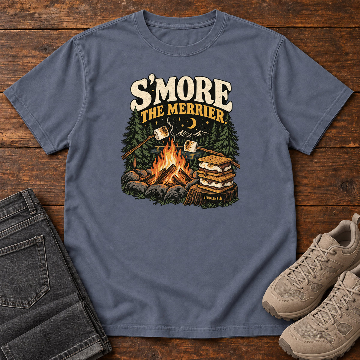 Denim Smore the Merrier T-Shirt with a campfire, marshmallows on sticks and stacked s’mores beneath a crescent moon; front flat lay on wood.