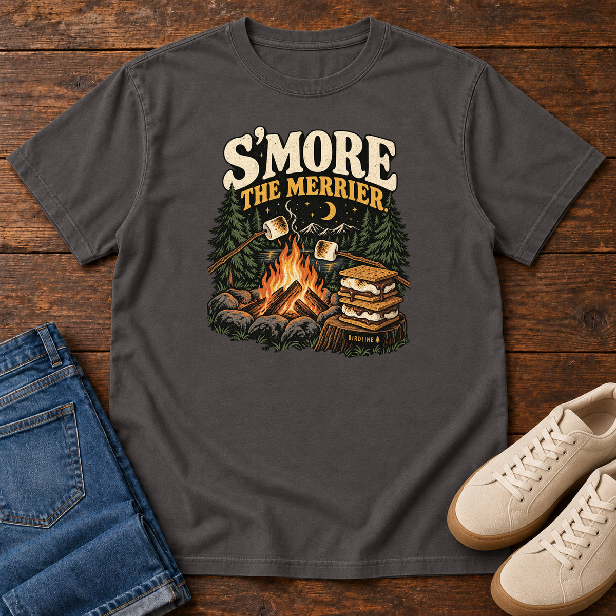Graphite Smore the Merrier T-Shirt with a campfire, marshmallows on sticks and stacked s’mores beneath a crescent moon; front flat lay on wood.