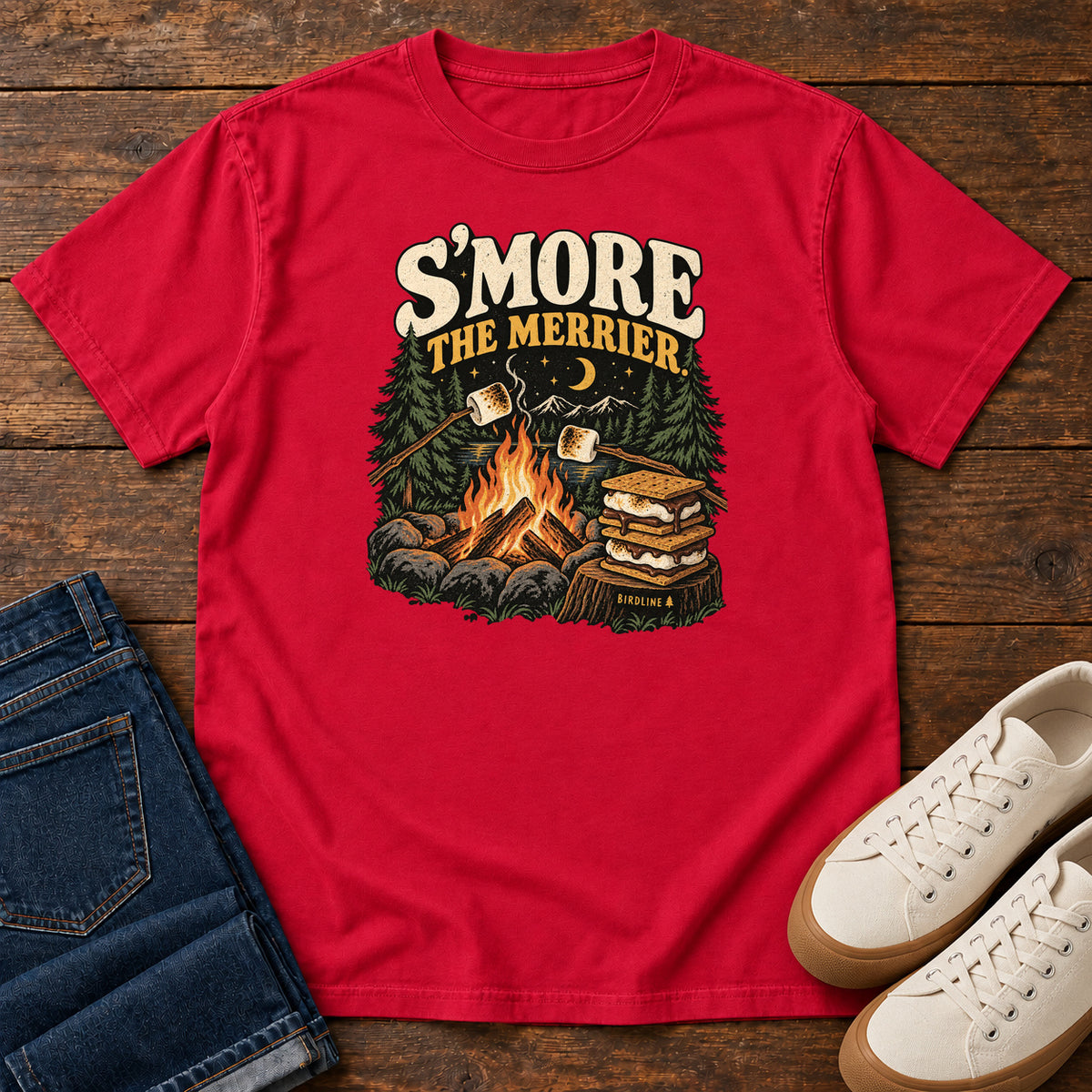 Red Smore the Merrier T-Shirt with a campfire, marshmallows on sticks and stacked s’mores beneath a crescent moon; front flat lay on wood.