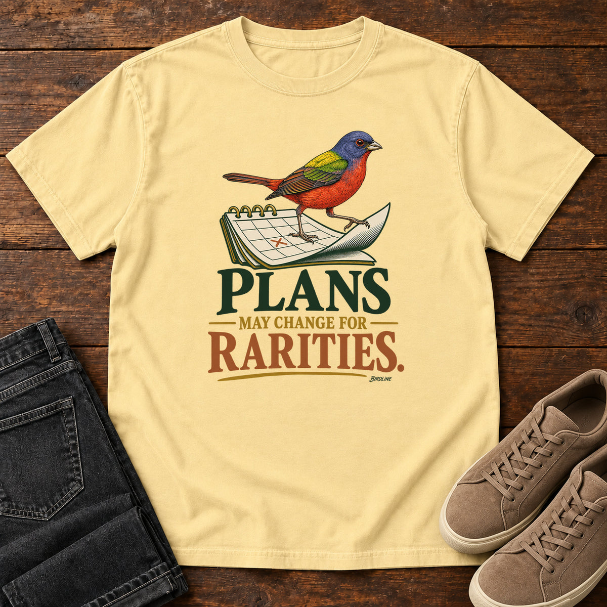 Butter Plans May Change for Rarities T-Shirt with a red, blue and green bird perched on a curled calendar page; front flat lay on wood.