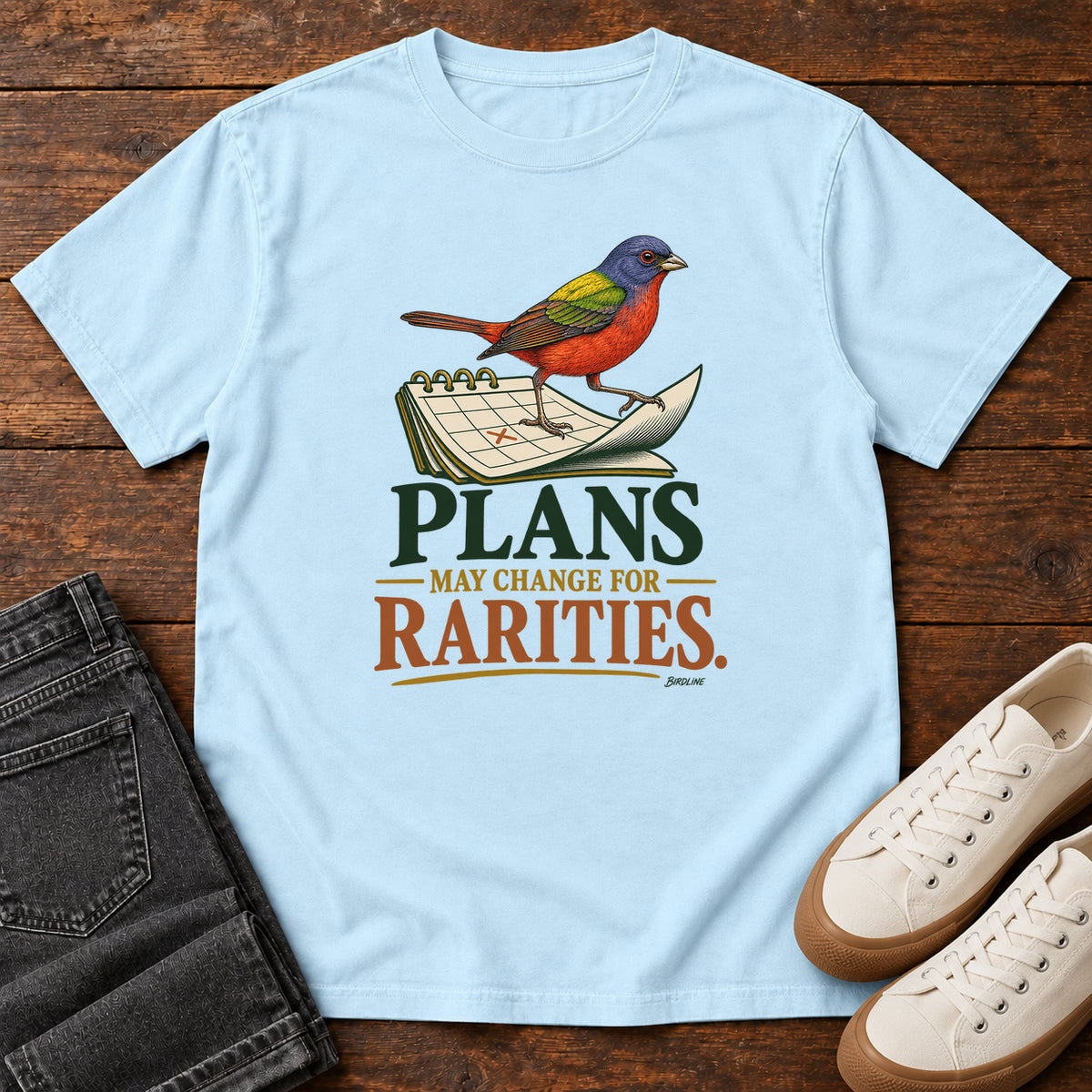 Chambray Plans May Change for Rarities T-Shirt with a red, blue and green bird perched on a curled calendar page; front flat lay on wood.