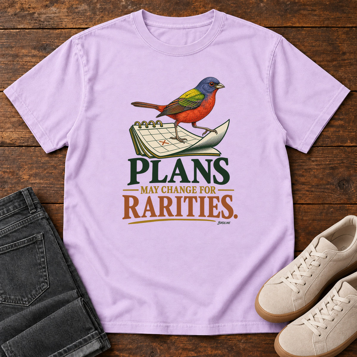 Orchid Plans May Change for Rarities T-Shirt with a red, blue and green bird perched on a curled calendar page; front flat lay on wood.