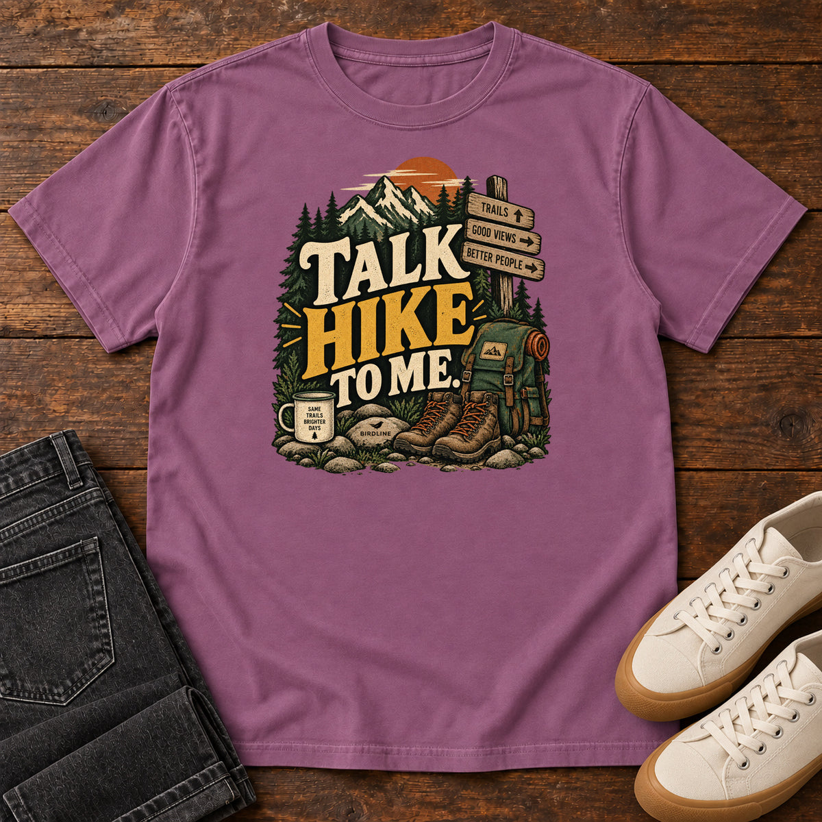 Berry Talk Hike to Me T-Shirt with hiking boots, a backpack and trail sign beneath mountains and bold lettering; front flat lay on wood.