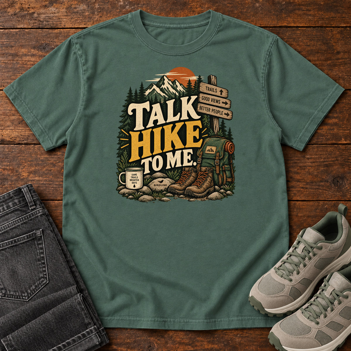 Blue Spruce Talk Hike to Me T-Shirt with hiking boots, a backpack and trail sign beneath mountains and bold lettering; front flat lay on wood.