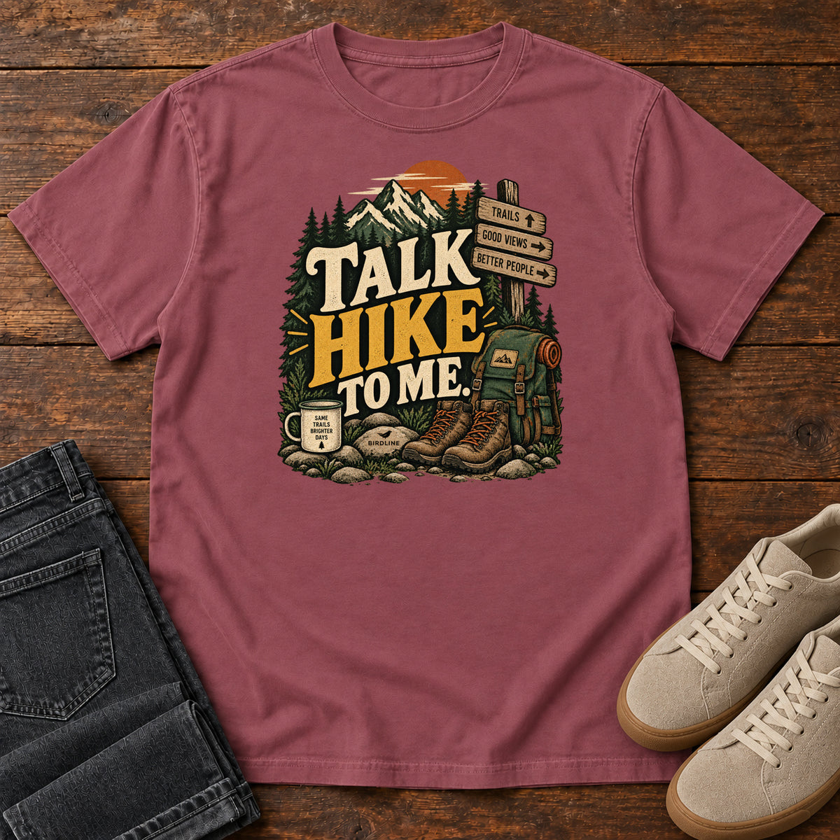 Brick Talk Hike to Me T-Shirt with hiking boots, a backpack and trail sign beneath mountains and bold lettering; front flat lay on wood.