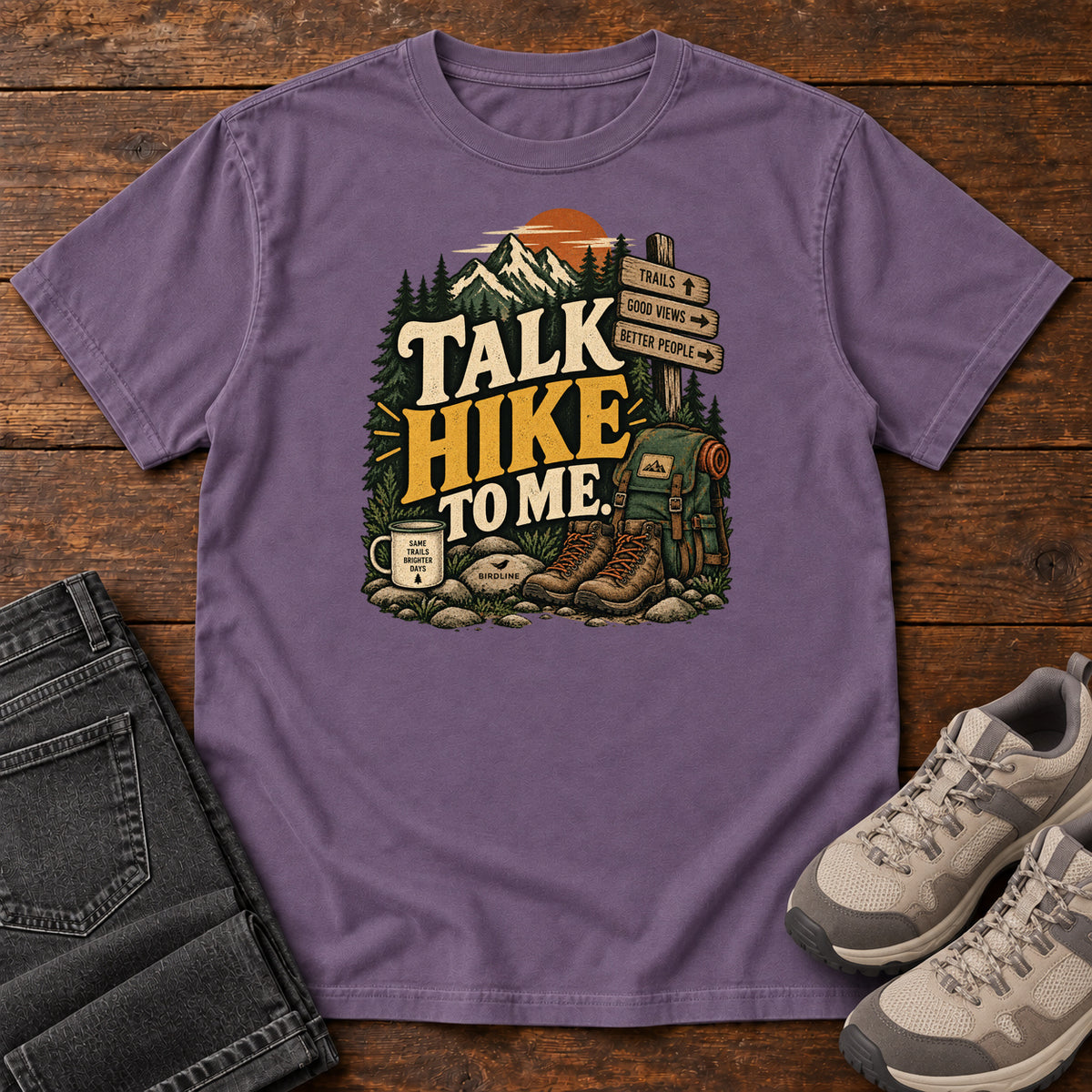 Grape Talk Hike to Me T-Shirt with hiking boots, a backpack and trail sign beneath mountains and bold lettering; front flat lay on wood.