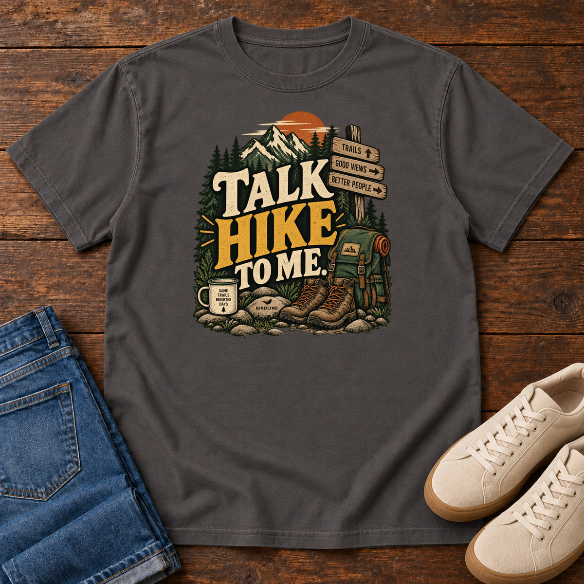 Graphite Talk Hike to Me T-Shirt with hiking boots, a backpack and trail sign beneath mountains and bold lettering; front flat lay on wood.