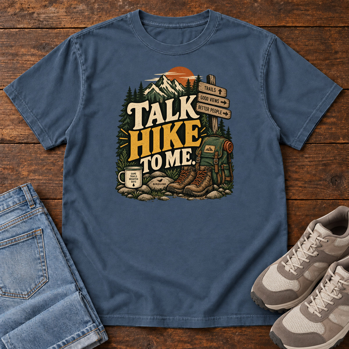 Midnight Talk Hike to Me T-Shirt with hiking boots, a backpack and trail sign beneath mountains and bold lettering; front flat lay on wood.