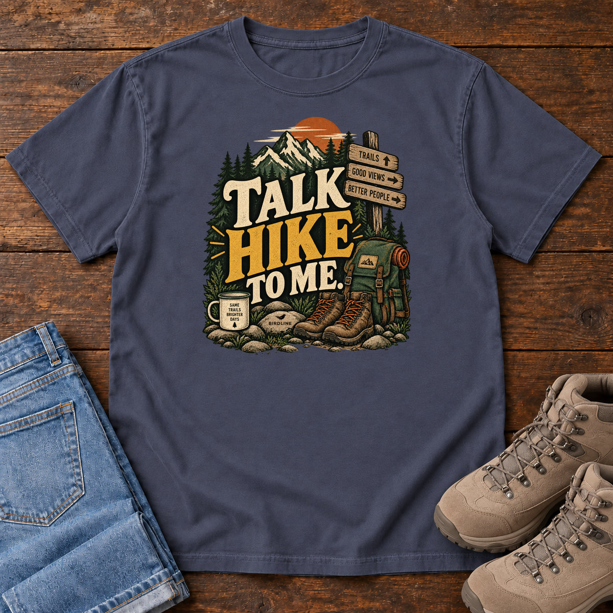 Navy Talk Hike to Me T-Shirt with hiking boots, a backpack and trail sign beneath mountains and bold lettering; front flat lay on wood.