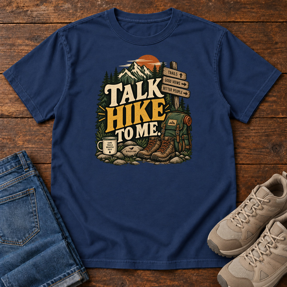 True Navy Talk Hike to Me T-Shirt with hiking boots, a backpack and trail sign beneath mountains and bold lettering; front flat lay on wood.