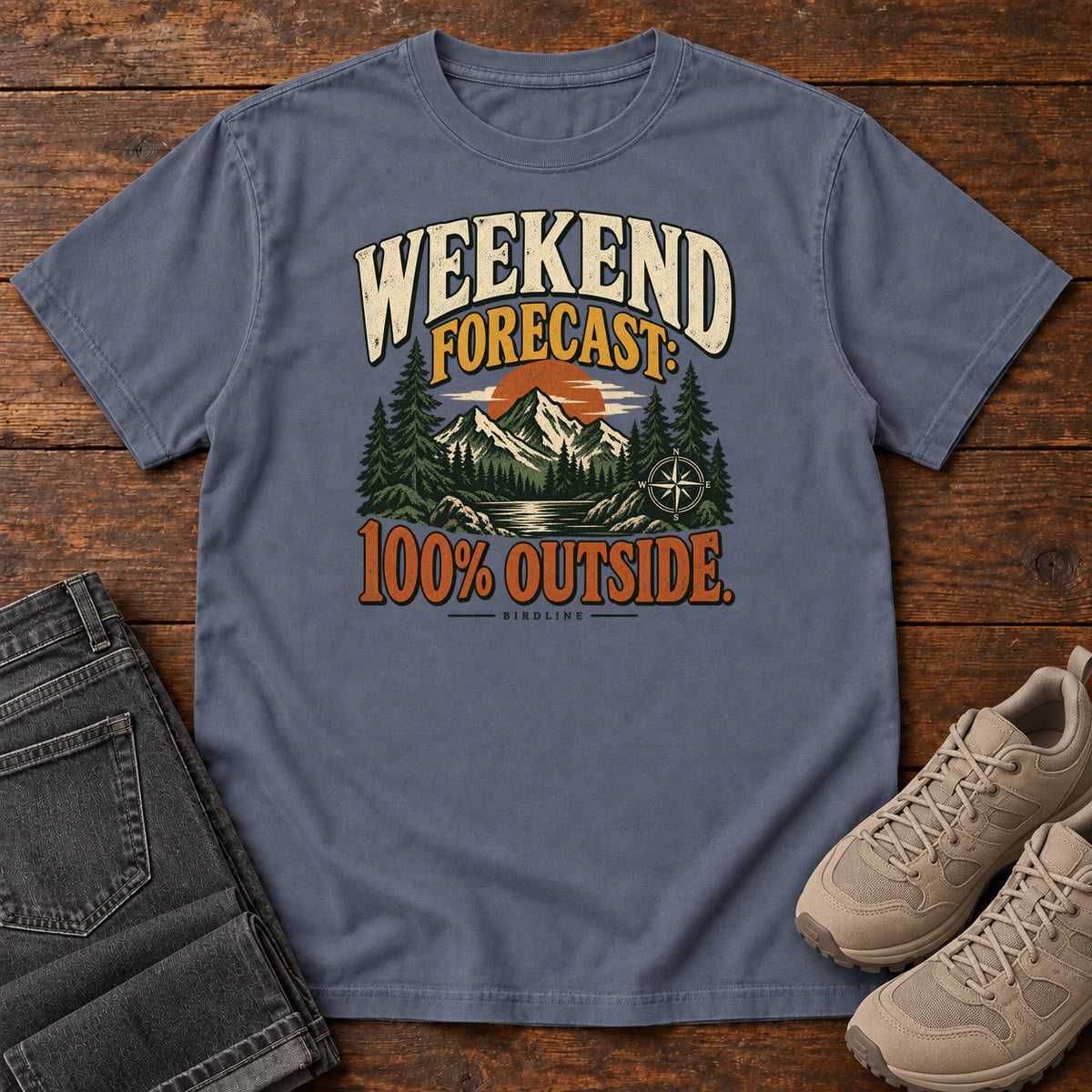 Denim Weekend Forecast 100 Percent Outside T-Shirt with mountains, evergreens, an orange sun and a compass beneath bold lettering; front flat lay on wood.