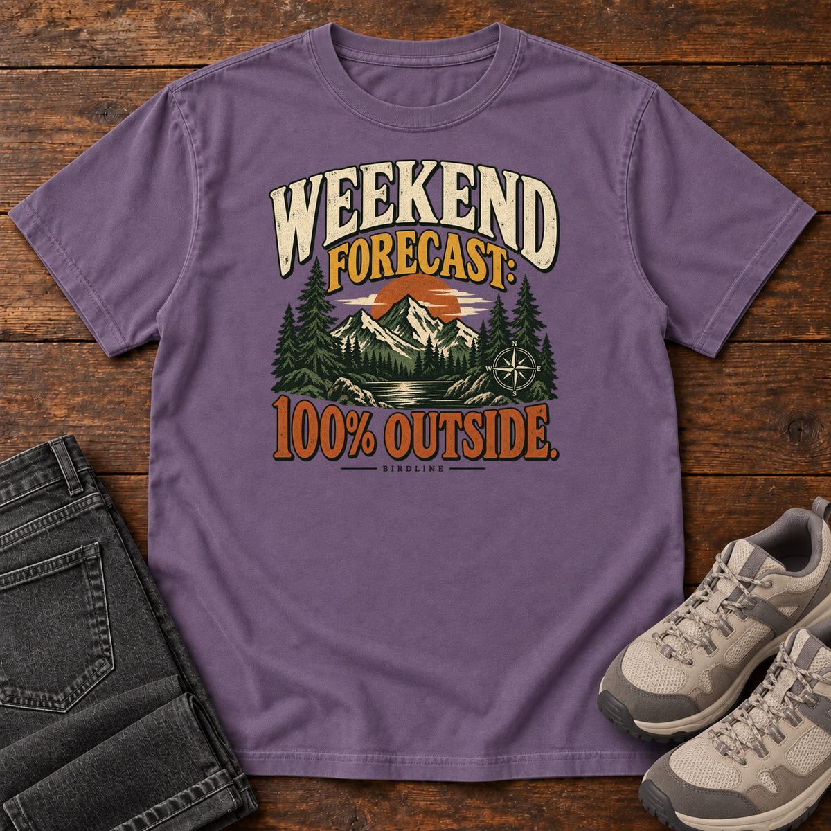 Grape Weekend Forecast 100 Percent Outside T-Shirt with mountains, evergreens, an orange sun and a compass beneath bold lettering; front flat lay on wood.