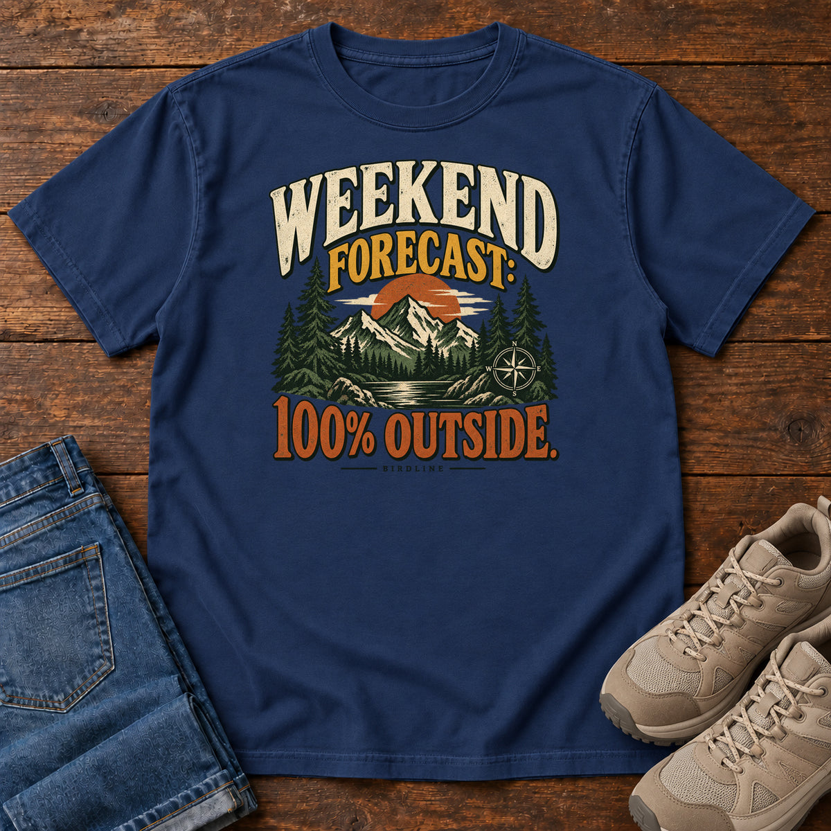 True Navy Weekend Forecast 100 Percent Outside T-Shirt with mountains, evergreens, an orange sun and a compass beneath bold lettering; front flat lay on wood.