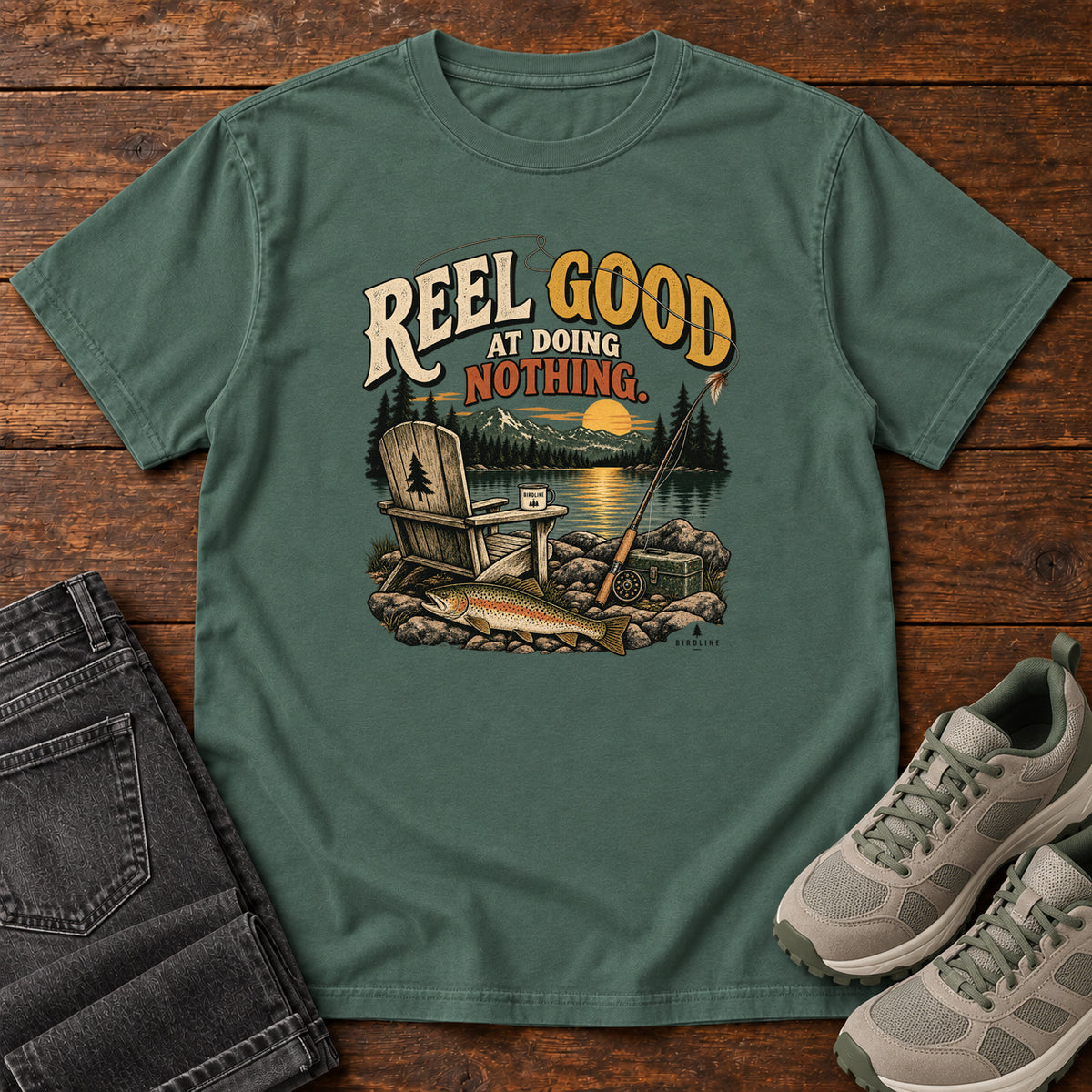 Blue Spruce Reel Good at Doing Nothing T-Shirt with a wooden chair and fishing rod beside a lake at sunset, front flat lay on wood.
