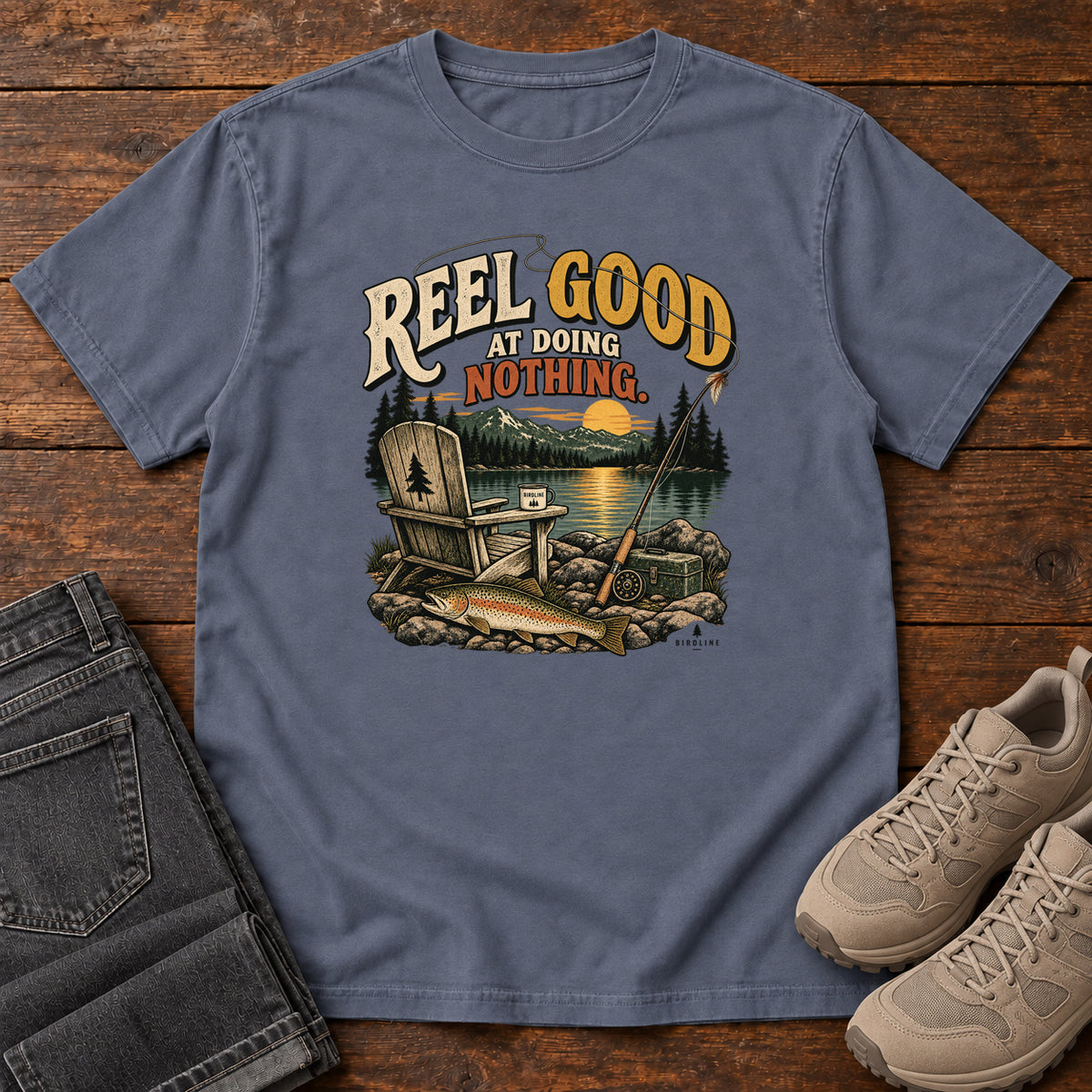 Denim Reel Good at Doing Nothing T-Shirt with a wooden chair and fishing rod beside a lake at sunset, front flat lay on wood.