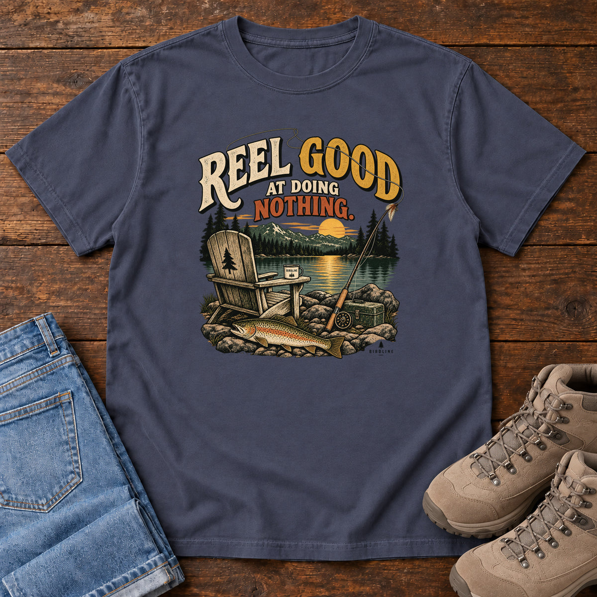 Navy Reel Good at Doing Nothing T-Shirt with a wooden chair and fishing rod beside a lake at sunset, front flat lay on wood.
