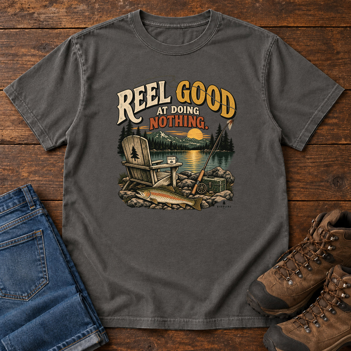 Pepper Reel Good at Doing Nothing T-Shirt with a wooden chair and fishing rod beside a lake at sunset, front flat lay on wood.