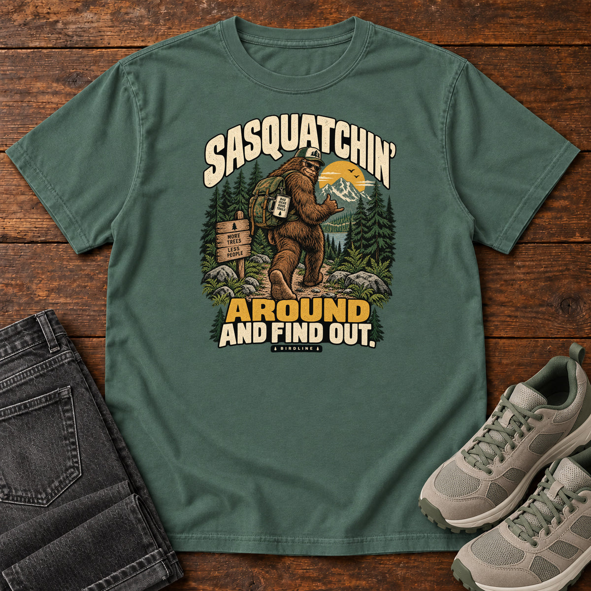 Blue Spruce Sasquatchin Around and Find Out T-Shirt with a backpack-wearing Sasquatch on a forest trail with cream-and-gold lettering, front flat lay on wood.