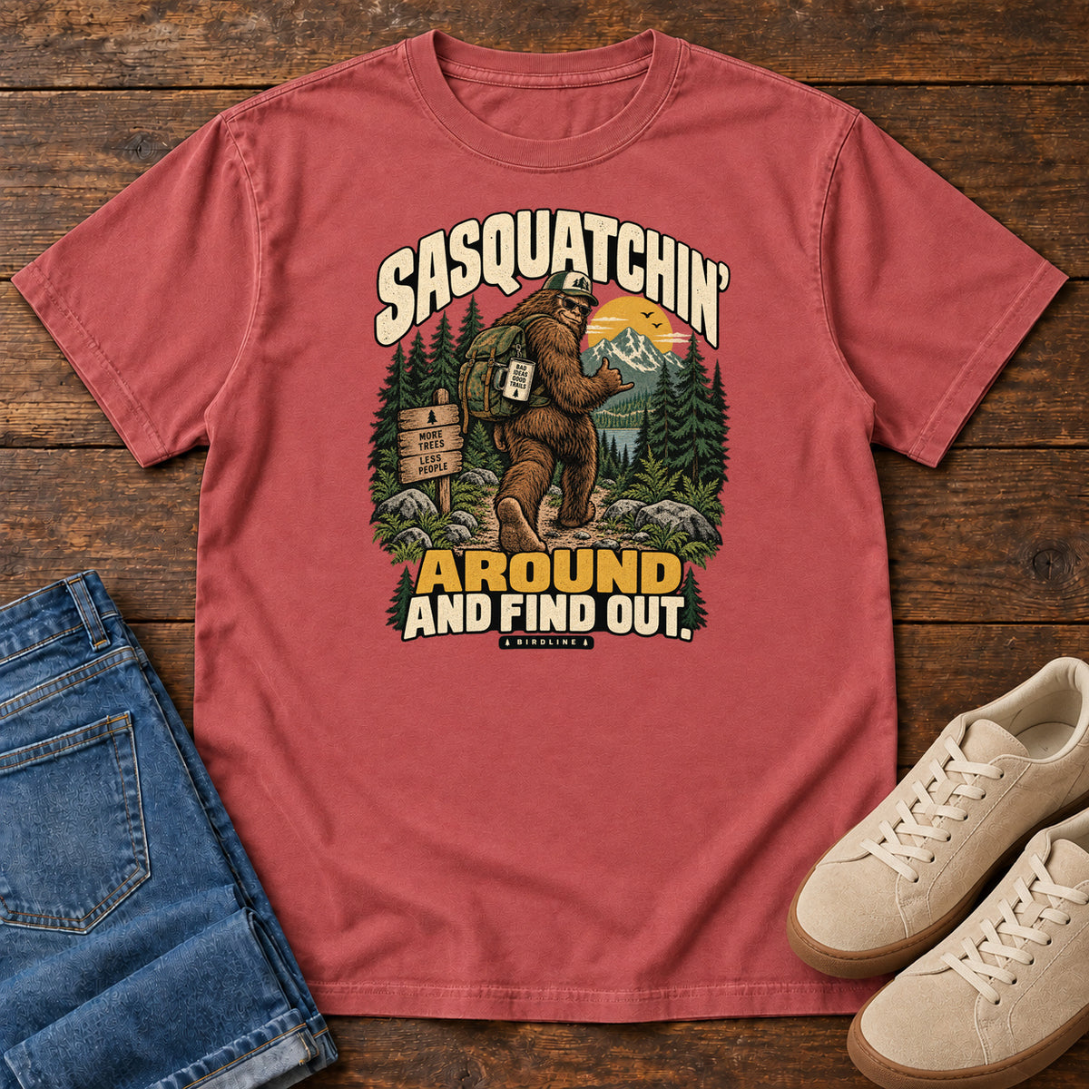 Crimson Sasquatchin Around and Find Out T-Shirt with a backpack-wearing Sasquatch on a forest trail with cream-and-gold lettering, front flat lay on wood.