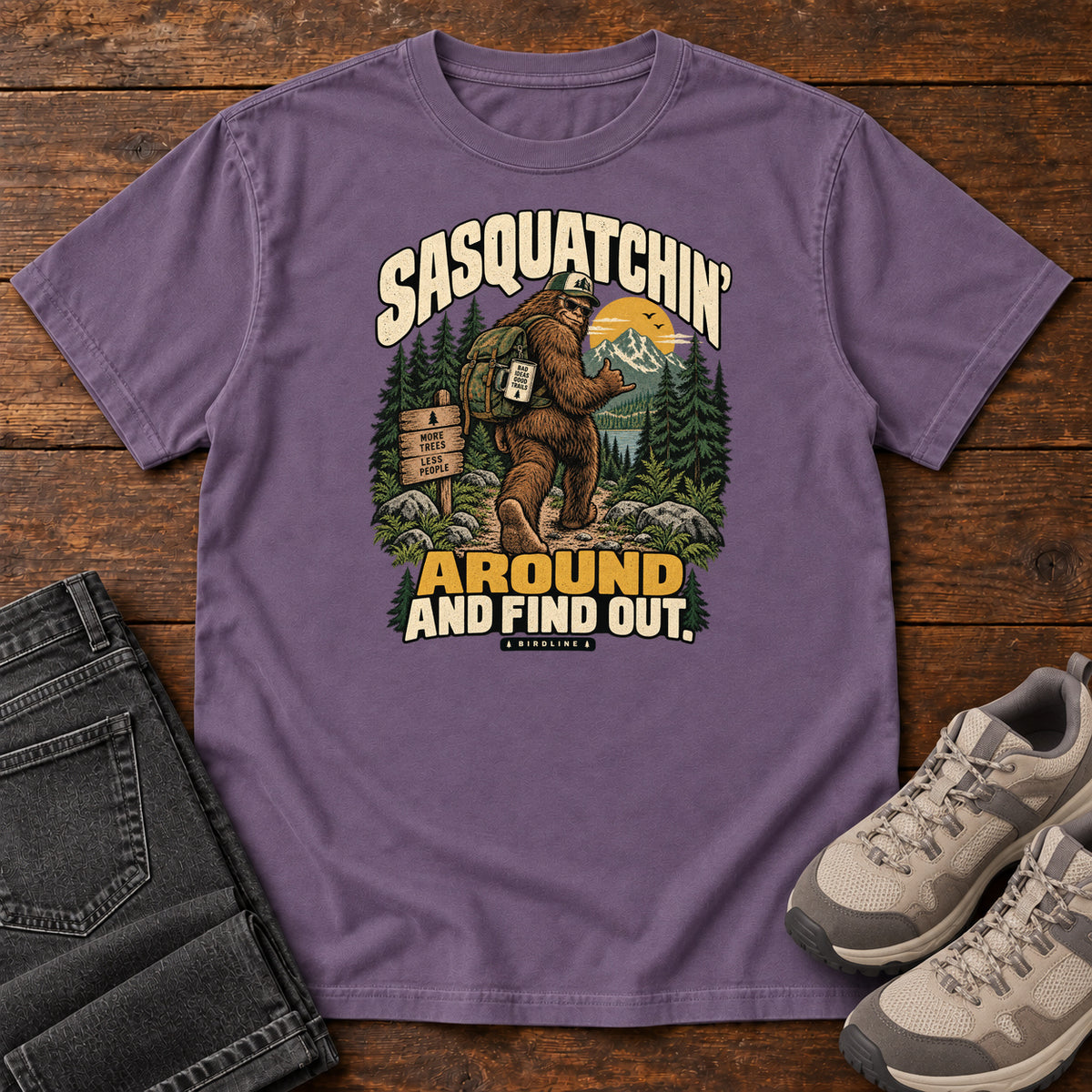 Grape Sasquatchin Around and Find Out T-Shirt with a backpack-wearing Sasquatch on a forest trail with cream-and-gold lettering, front flat lay on wood.