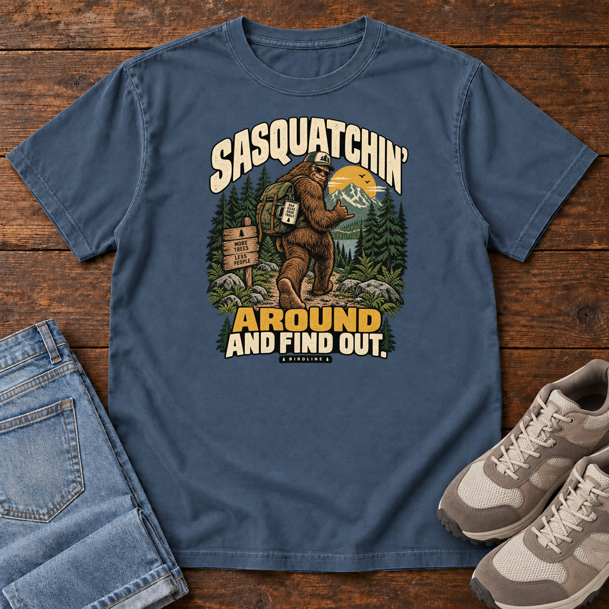 Midnight Sasquatchin Around and Find Out T-Shirt with a backpack-wearing Sasquatch on a forest trail with cream-and-gold lettering, front flat lay on wood.