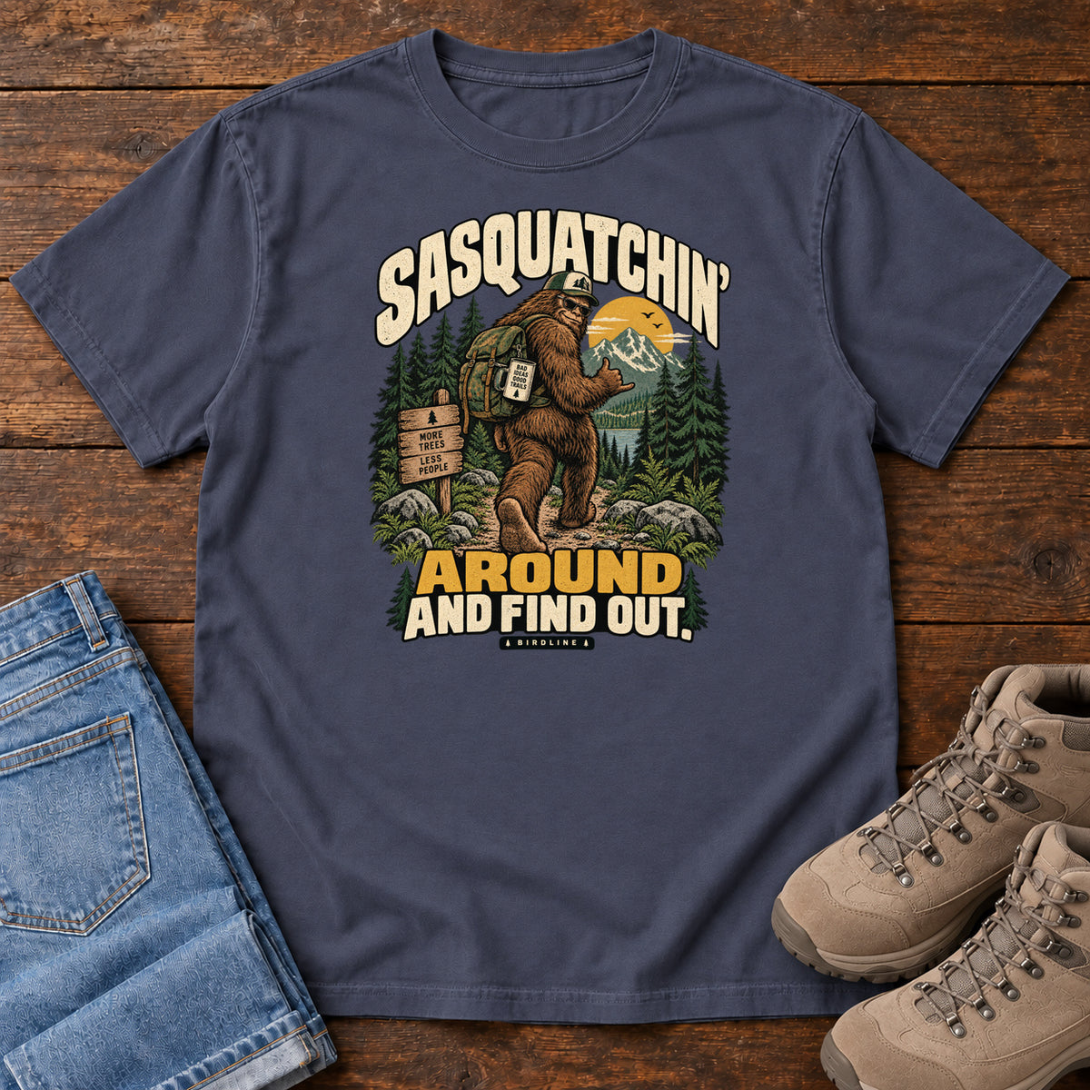 Navy Sasquatchin Around and Find Out T-Shirt with a backpack-wearing Sasquatch on a forest trail with cream-and-gold lettering, front flat lay on wood.