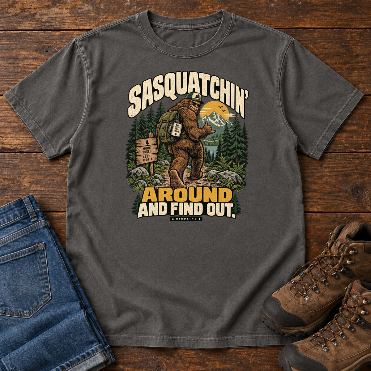 Pepper Sasquatchin Around and Find Out T-Shirt with a backpack-wearing Sasquatch on a forest trail with cream-and-gold lettering, front flat lay on wood.