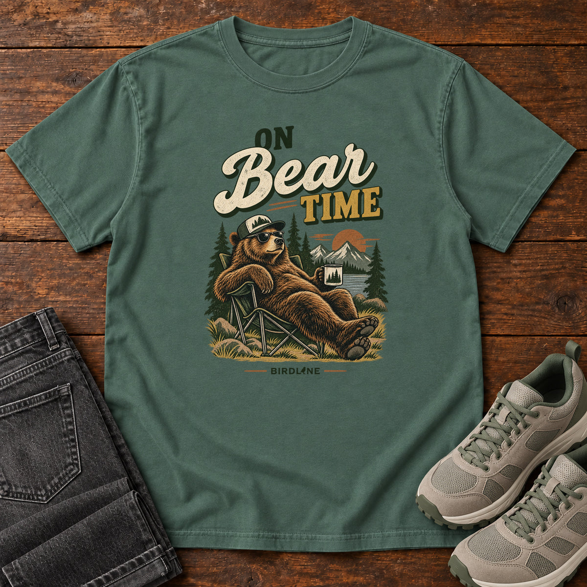 Blue Spruce On Bear Time T-Shirt with a bear in a cap and sunglasses reclining in a camp chair with a mug; front flat lay on wood.