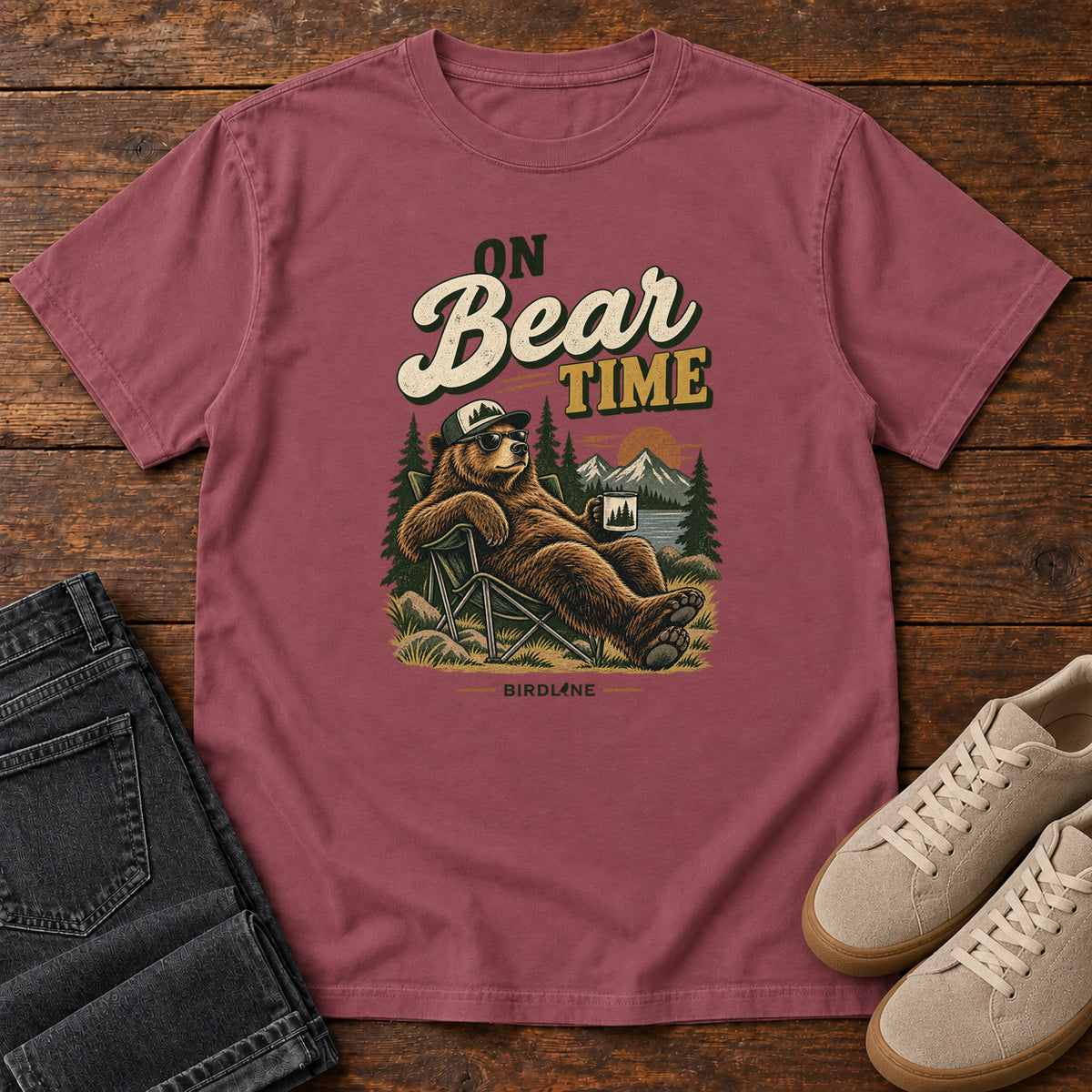 Brick On Bear Time T-Shirt with a bear in a cap and sunglasses reclining in a camp chair with a mug; front flat lay on wood.
