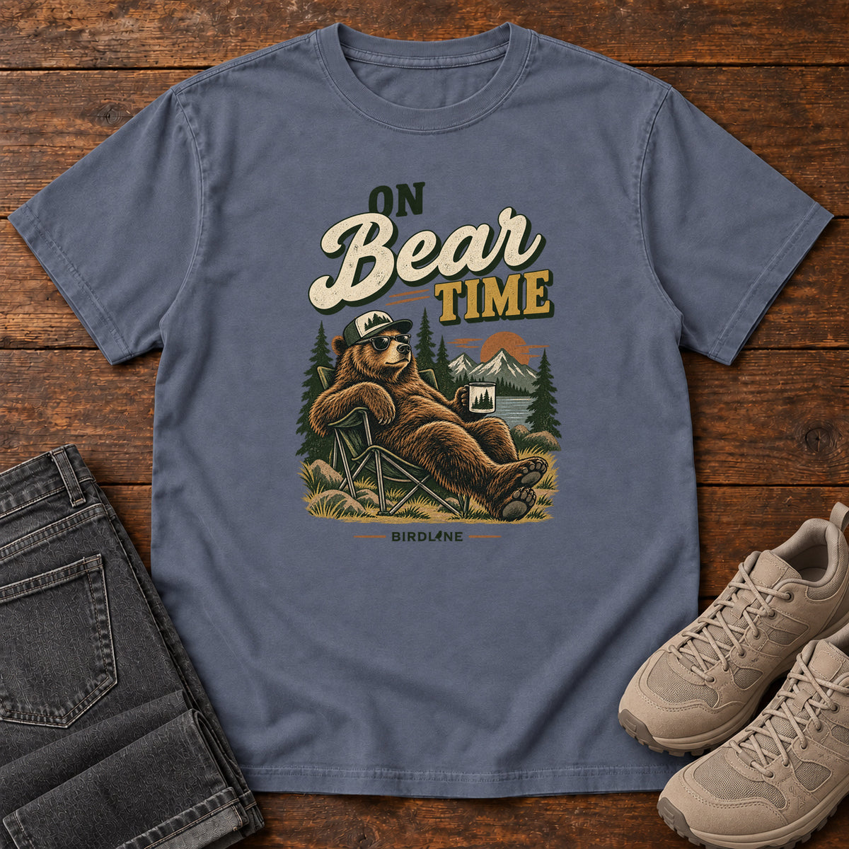 Denim On Bear Time T-Shirt with a bear in a cap and sunglasses reclining in a camp chair with a mug; front flat lay on wood.