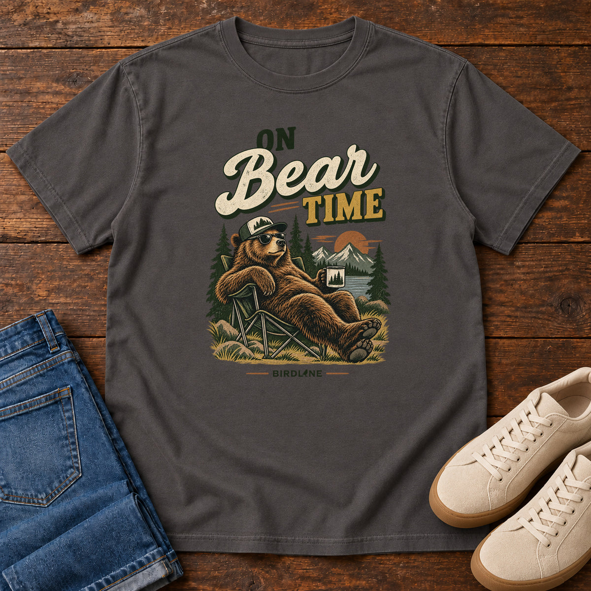 Graphite On Bear Time T-Shirt with a bear in a cap and sunglasses reclining in a camp chair with a mug; front flat lay on wood.