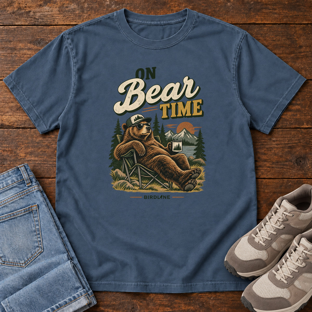 Midnight On Bear Time T-Shirt with a bear in a cap and sunglasses reclining in a camp chair with a mug; front flat lay on wood.