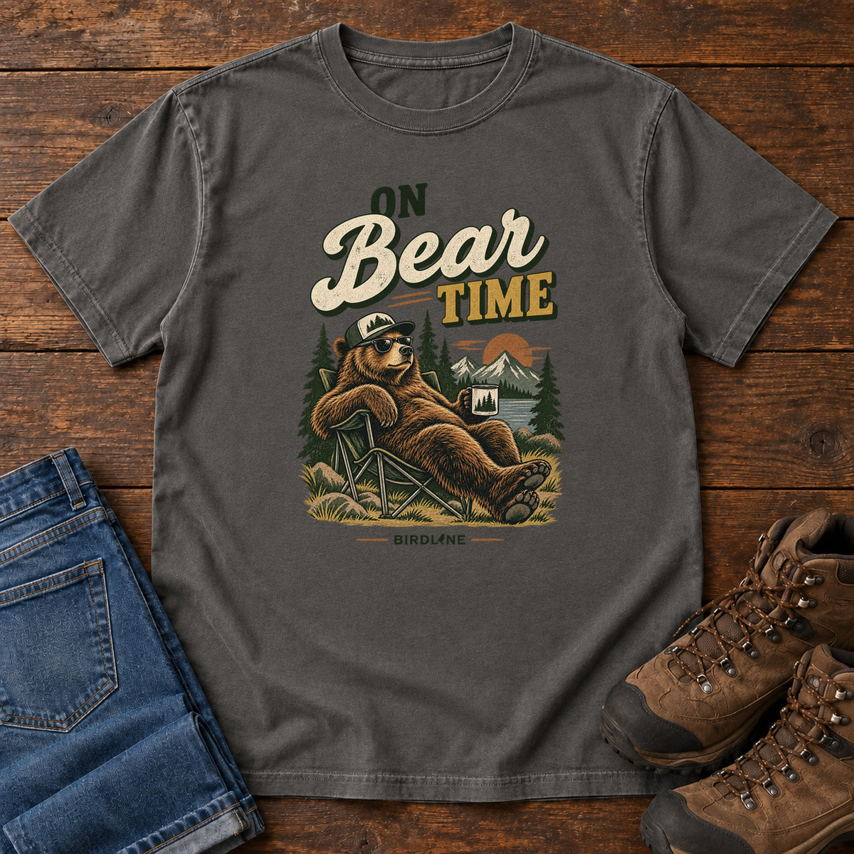 Pepper On Bear Time T-Shirt with a bear in a cap and sunglasses reclining in a camp chair with a mug; front flat lay on wood.