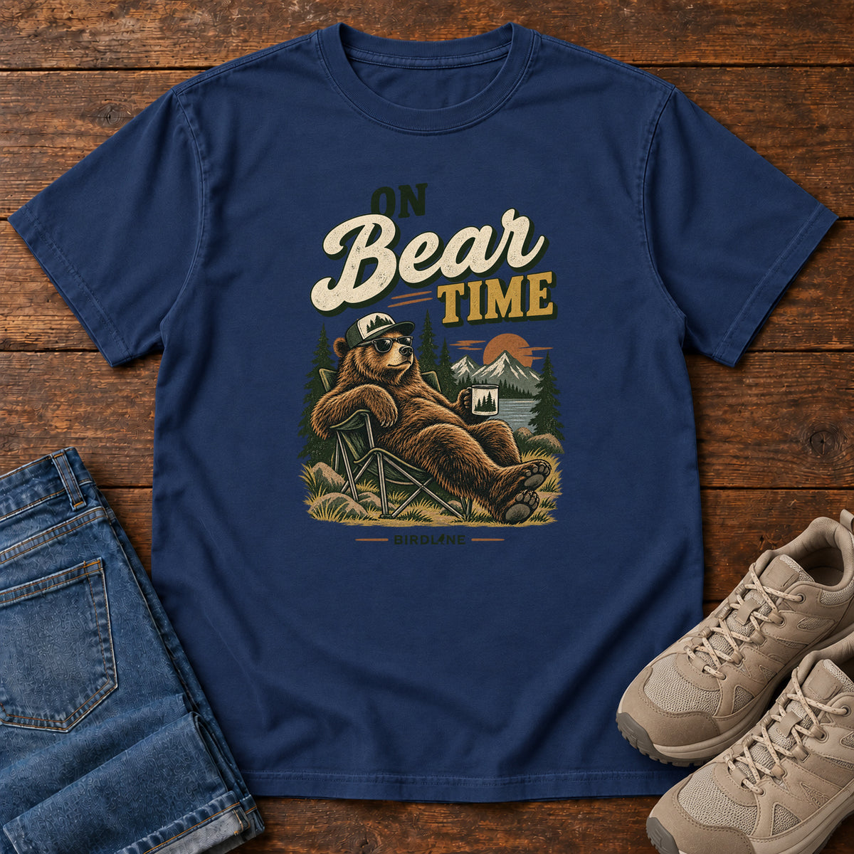 True Navy On Bear Time T-Shirt with a bear in a cap and sunglasses reclining in a camp chair with a mug; front flat lay on wood.