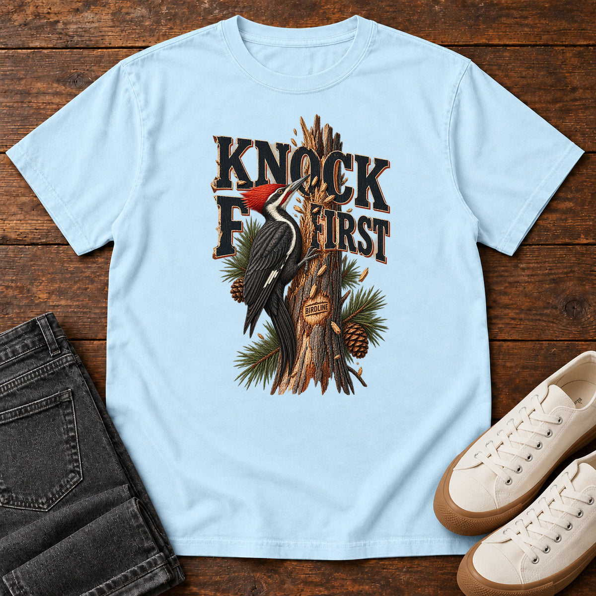 Chambray Knock First Woodpecker T-Shirt with a red-crested woodpecker on a pine trunk beneath Knock First lettering, front flat lay on wood.