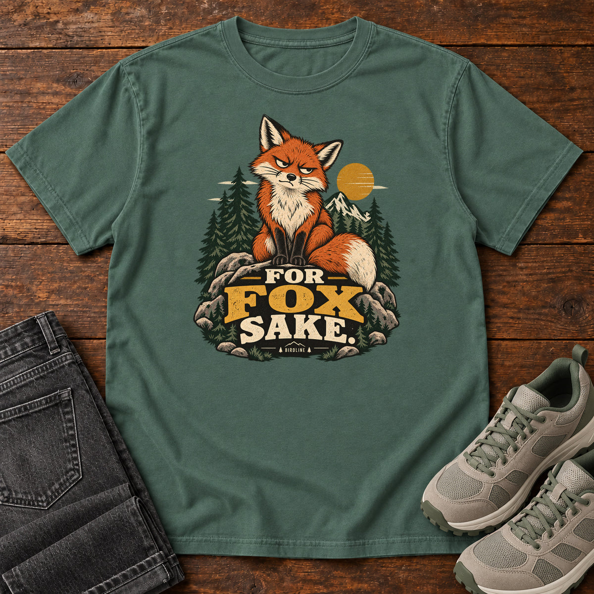 Blue Spruce For Fox Sake T-Shirt with an orange fox sitting on rocks against pines, mountains and a golden sun; front flat lay on wood.