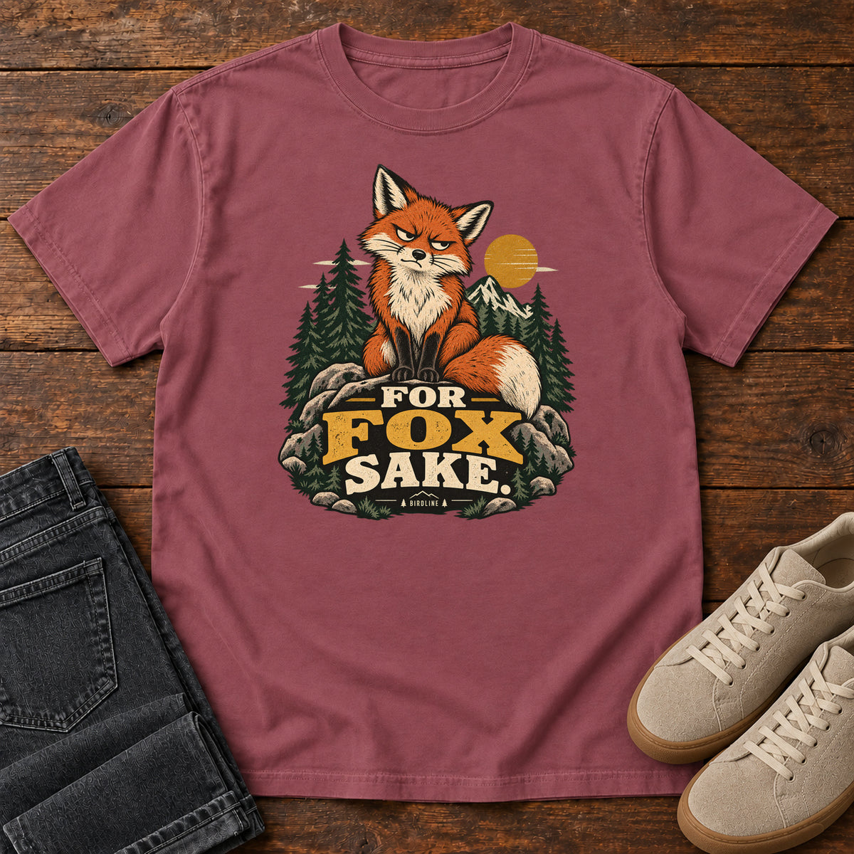 Brick For Fox Sake T-Shirt with an orange fox sitting on rocks against pines, mountains and a golden sun; front flat lay on wood.