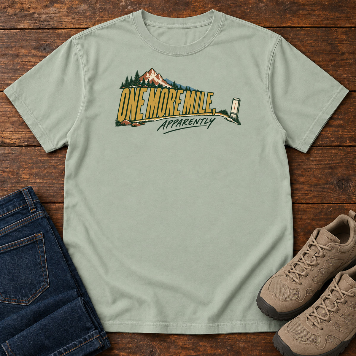 One More Mile Apparently T-Shirt in Bay, front laid flat on wood, featuring gold slogan lettering with a mountain, pine trees and a small trail marker.